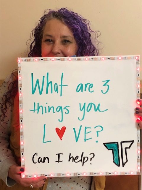 Shawna Hugill holds up a sign in the hallway that reads, What are 3 things you love? Can I help?